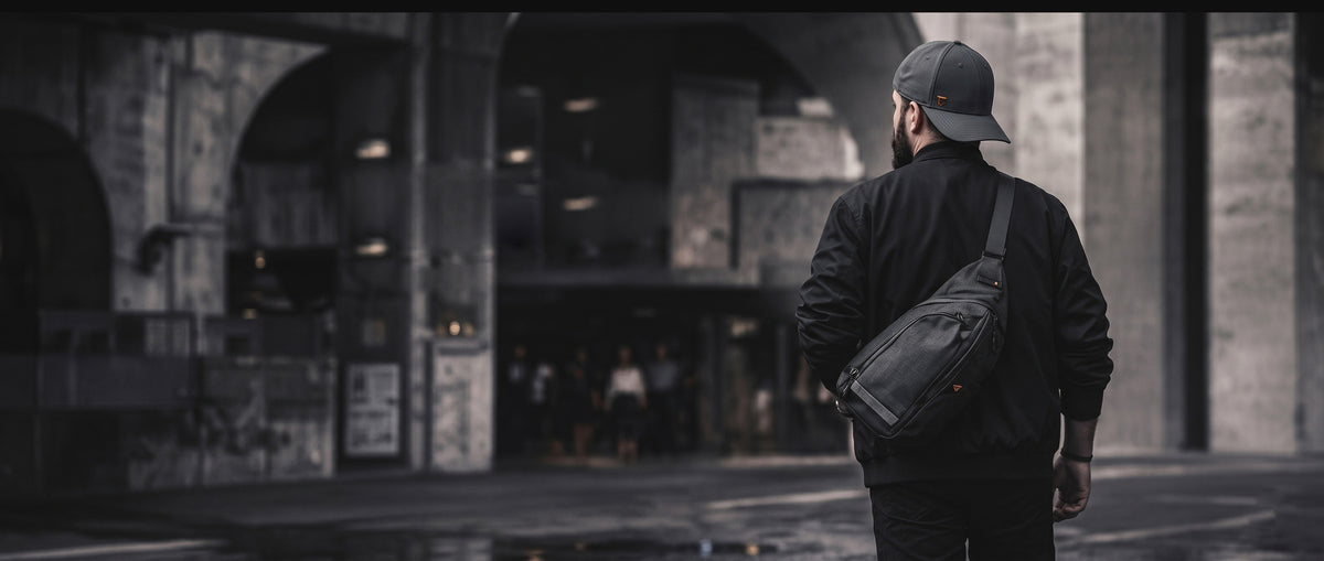 Rear view of a man walking in a moody urban city setting, wearing a luxury dark grey cap with a subtle orange logo. He is styled in premium dark streetwear, featuring a black jacket and a crossbody bag, captured with high-quality cinematic lighting.
